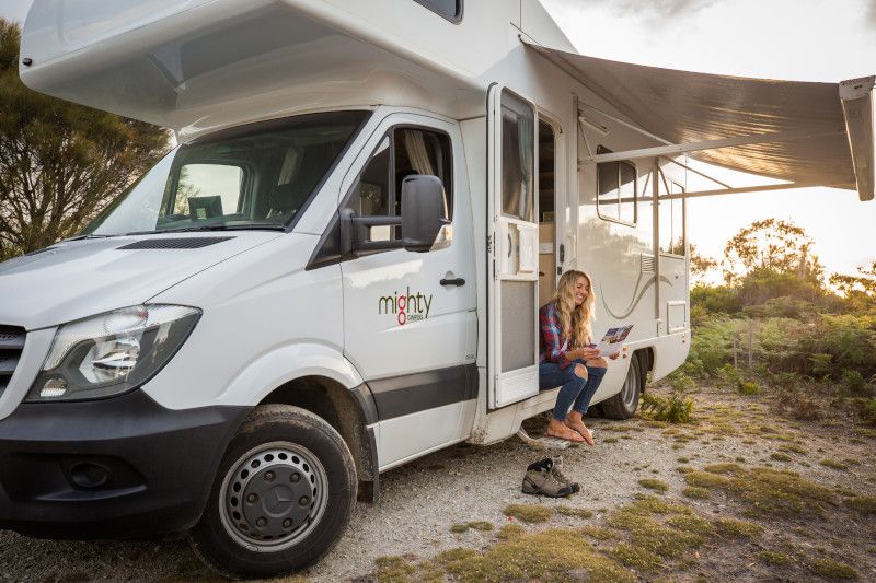 A woman sitting in the doorway of a parked white campervan with its awning extended, reading a map.