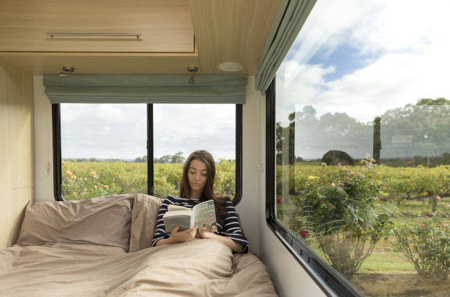 A woman reads a book in a campervan bed, with large windows overlooking a green rose garden.