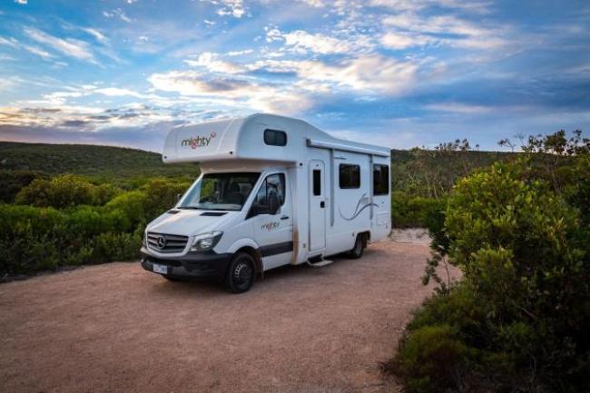 White Mighty campervan parked on a dirt track surrounded by green bushes under a cloudy blue sky.