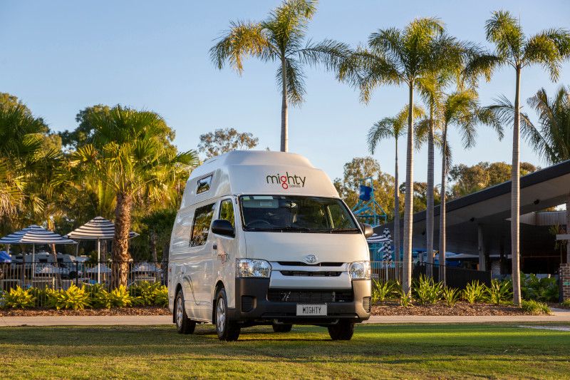 White Mighty campervan parked on a grassy lawn, surrounded by tall palm trees on a sunny day.