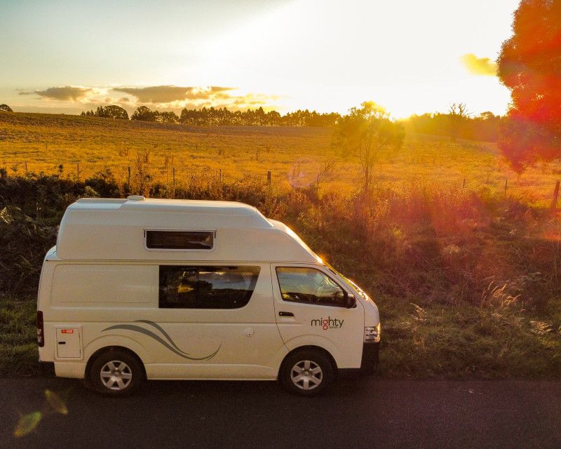 White Mighty campervan parked by a road next to a grassy field at sunset.
