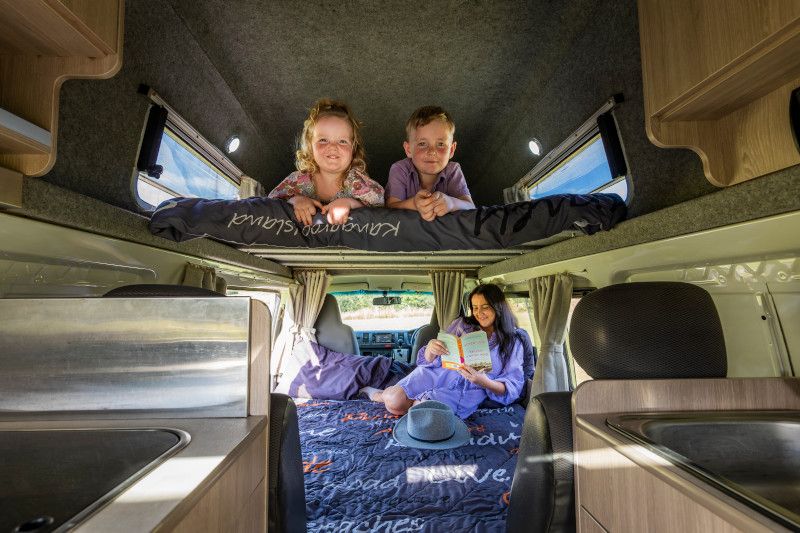 Two children smile from a campervan loft bed above a woman reading on the lower bed.