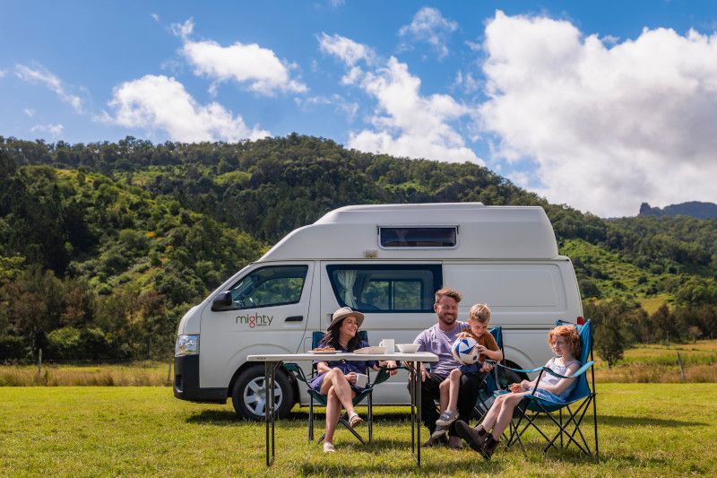 A family of four sits at a camping table next to a white campervan parked in a grassy field.