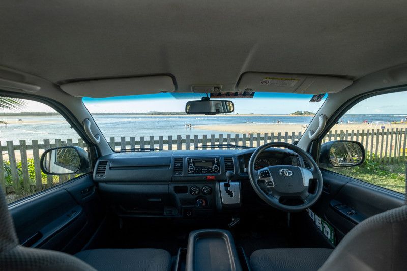 Interior view of a right-hand drive Toyota campervan looking out at a beach behind a timber fence.