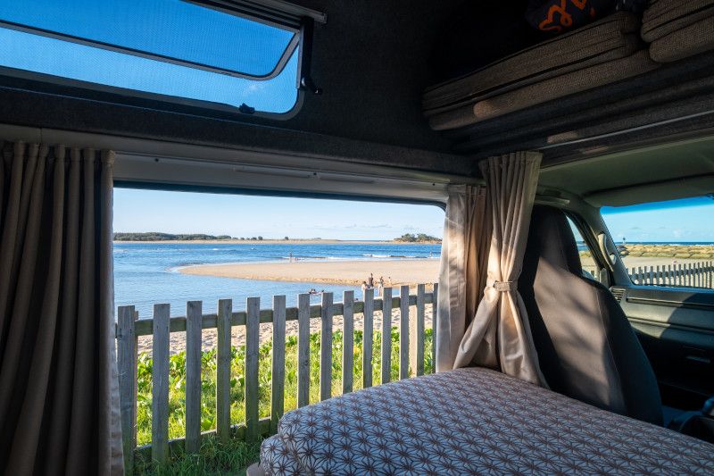 Inside a campervan, looking past a patterned bed towards a timber fence, sandy beach, and calm blue water.