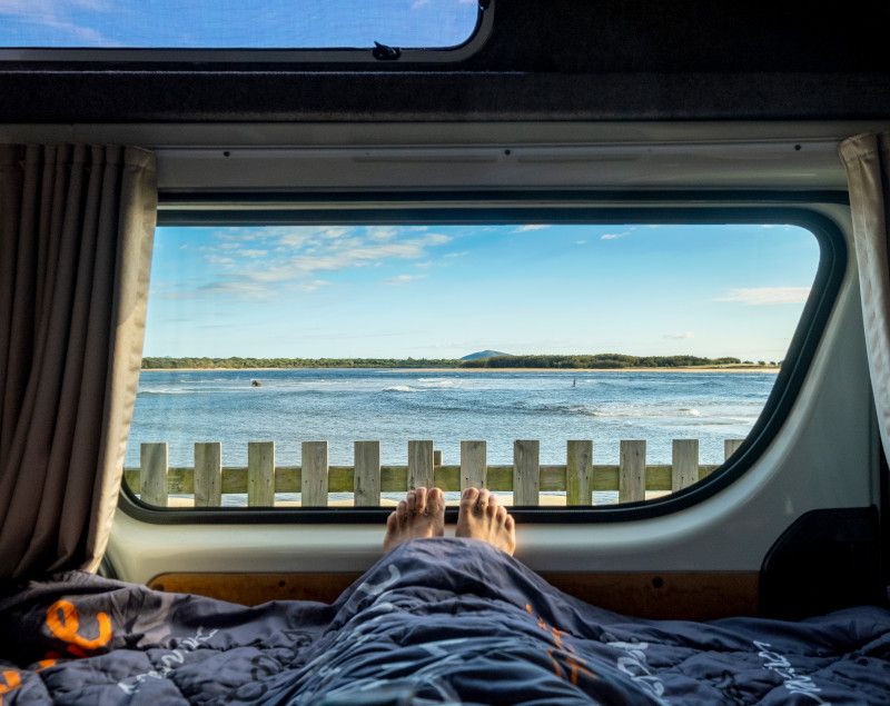 Feet sticking out from a sleeping bag in a campervan, looking through the window at a timber fence and the ocean.