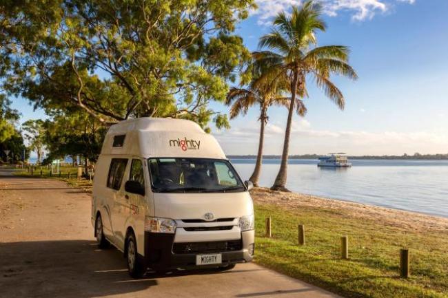 Mighty campervan parked on a path beside a sandy beach with palm trees and calm water.