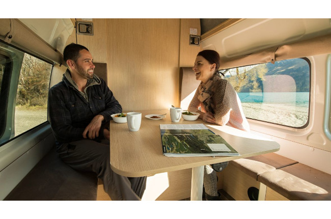 Man and woman sitting at a table inside a campervan, smiling with a lake and mountain view visible through the window.