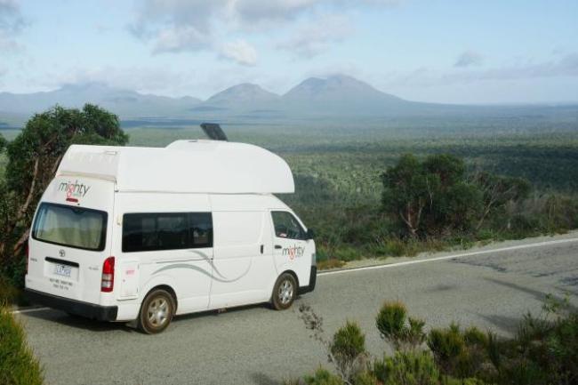 White Mighty campervan parked on a roadside lookout overlooking a vast forest landscape and mountains.