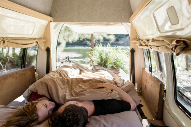 Couple lying in a campervan bed looking at each other, with open back doors revealing a leafy outdoor river view.