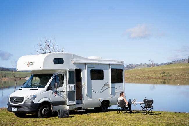 White Mighty campervan parked beside a lake with a person sitting on a camp chair looking at the water.