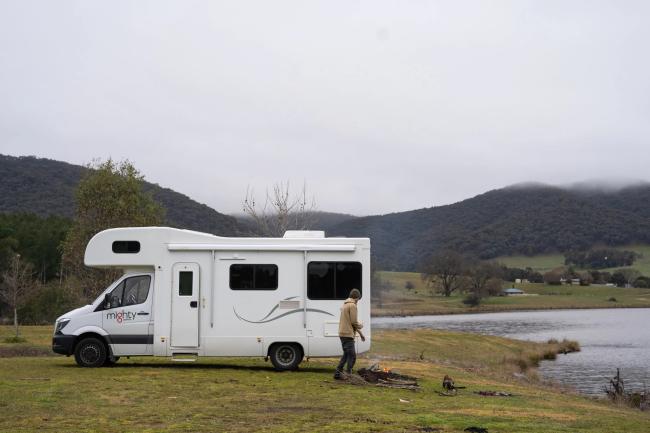 White campervan parked by a lake, with a person standing near a campfire against a backdrop of misty hills.