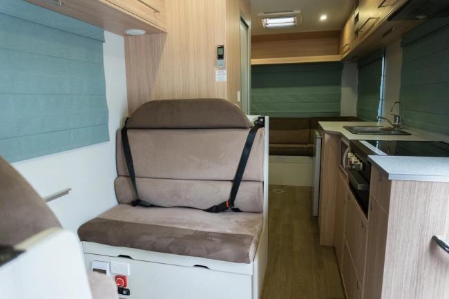 Campervan interior featuring a brown fabric seat with seatbelts, a light timber-look kitchenette, and blue roman blinds.