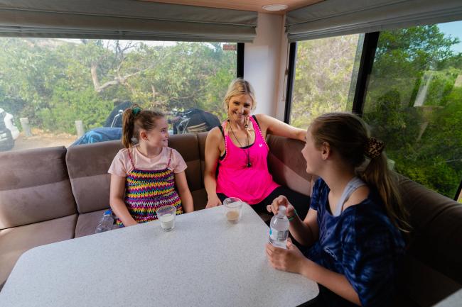 A woman and two young girls smiling and chatting at a dining table inside a campervan booth.