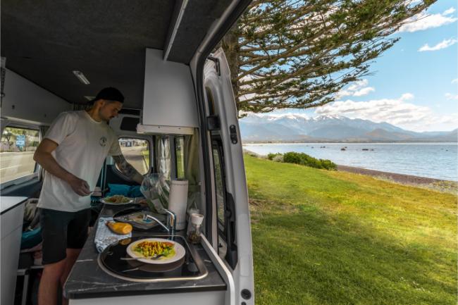 Man preparing food in a campervan kitchen with the side door open to a grassy shoreline and distant mountains.