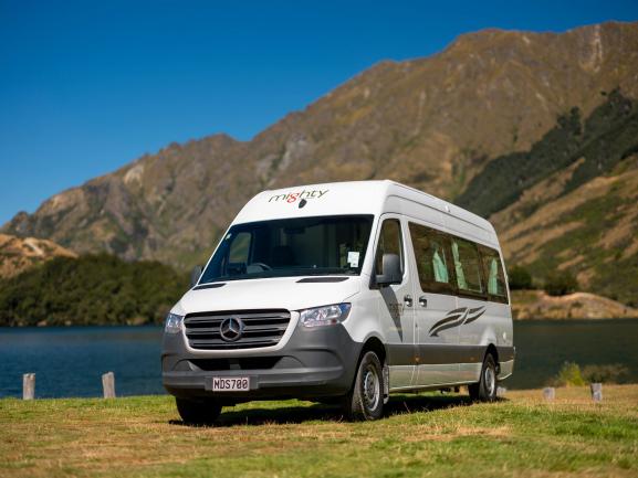 White Mercedes Mighty campervan parked on grass beside a lake with mountains in the background.