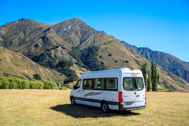 White campervan parked on a grassy field in front of dry, rugged mountains under a clear blue sky.