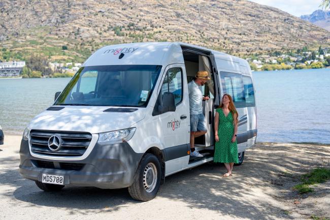 A man steps out of a white Mighty campervan parked by a lake as a woman in a green dress stands outside.