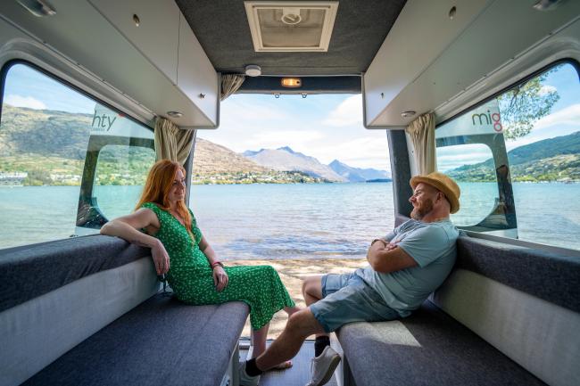 A man and woman relax inside a campervan, looking out the open back doors at a lake and mountains.