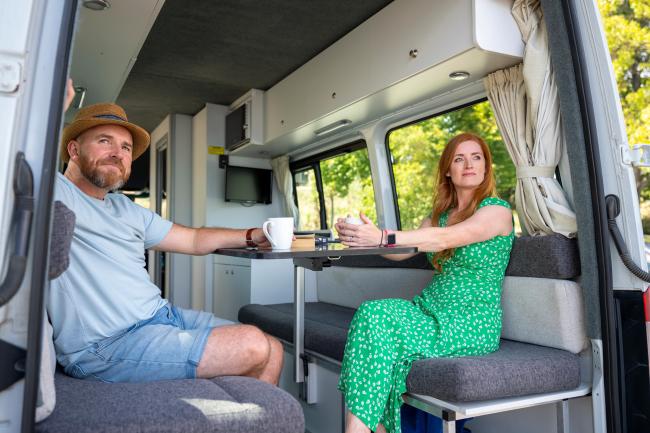 A man and a woman relax inside a modern campervan interior, sitting at a table with mugs.