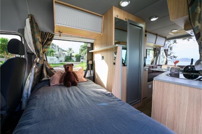Campervan interior featuring a bed with a teddy bear, light timber cabinetry, and a kitchen counter.