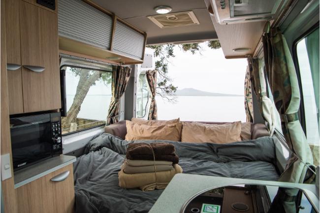 Interior of a campervan with a made bed, timber cabinets, and a sea view through the open back doors.