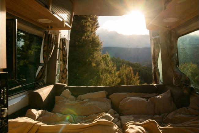 View from a campervan bed looking out open back doors at a sunlit forest and distant mountains.