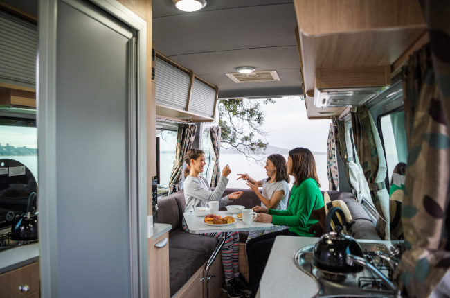 A woman and two girls play a hand game around a table inside a campervan, with scenic water views in the background.