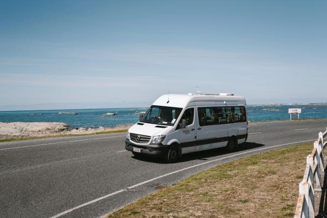 White Mercedes-Benz campervan travelling on a coastal road next to the ocean.
