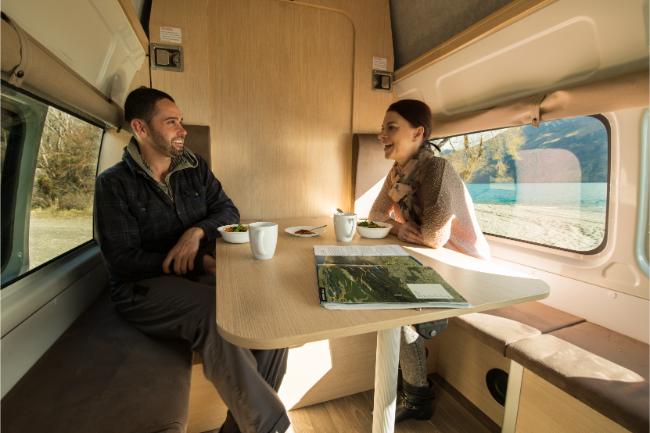 A man and woman sit at a table inside a campervan, smiling at each other with a scenic lake view through the window.