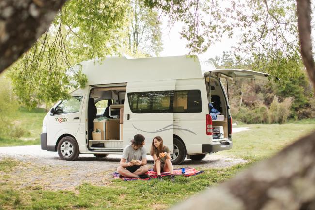 A couple picnics on a blanket next to a white campervan with open doors in a grassy, tree-lined clearing.