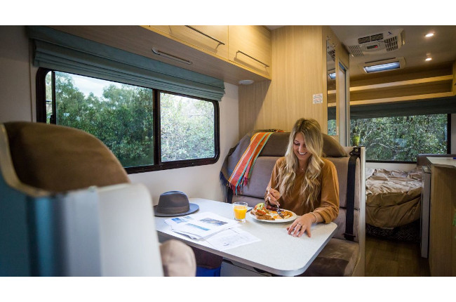 Woman eating breakfast at a dining booth inside a campervan with a window view of green trees.