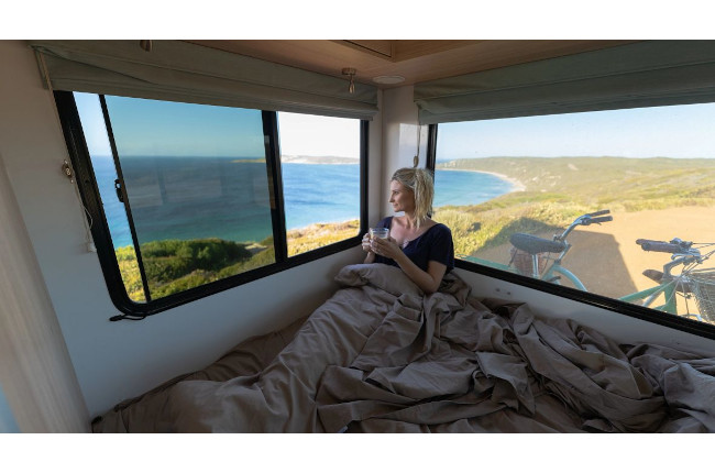 Woman inside a campervan bed holding a mug, looking out at a coastal ocean view with a bicycle outside.