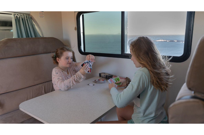 Two girls play a card game at a table inside a campervan with an ocean view visible through the window.