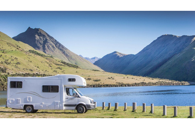 White campervan parked on a grassy bank beside a blue lake, with green mountains in the background.