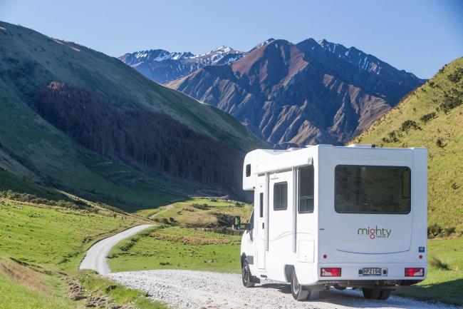 White campervan on a winding gravel road in a green valley with distant mountains.