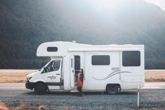 Woman standing in the doorway of a white Mighty campervan on a gravel roadside with mountains in the background.