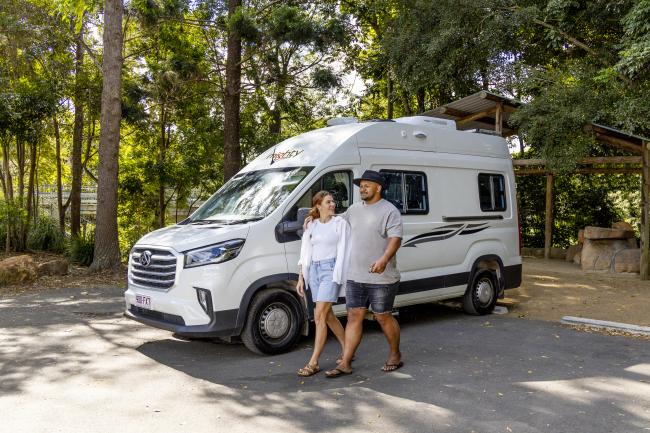 A couple walking past a white campervan parked in a shaded, tree-lined area.