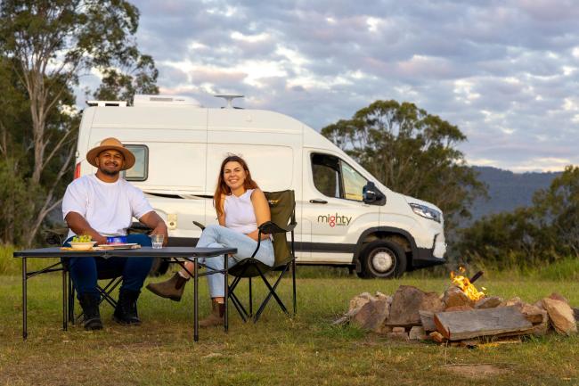 A man and woman relax next to a white Mighty campervan with a small campfire in the foreground.