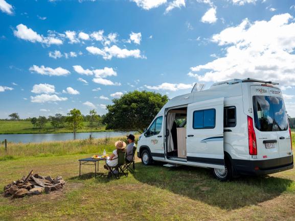 Couple sitting beside a white campervan overlooking a river, with a stone fire pit in the grassy foreground.