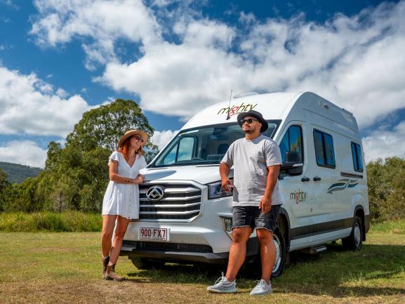 Couple posing in front of a white Mighty campervan in a grassy field under a blue sky.