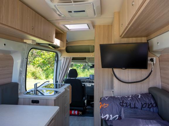 Campervan interior featuring a kitchen sink, wall-mounted TV, bed, and a walkthrough to the front cabin.
