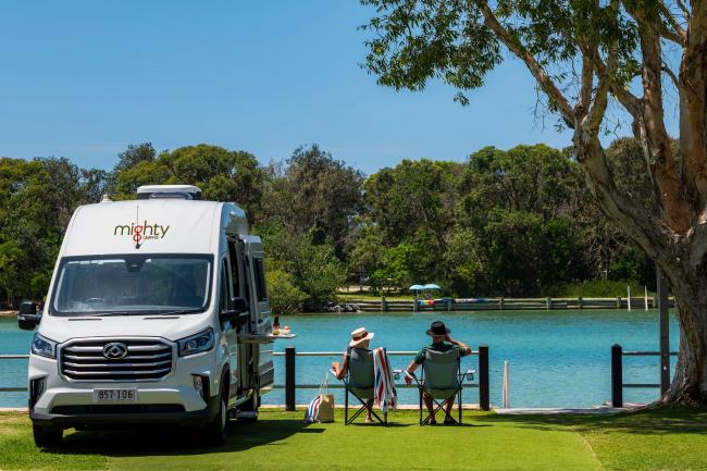 Two people in camp chairs relax by a river next to a parked white Mighty campervan on a grassy bank.