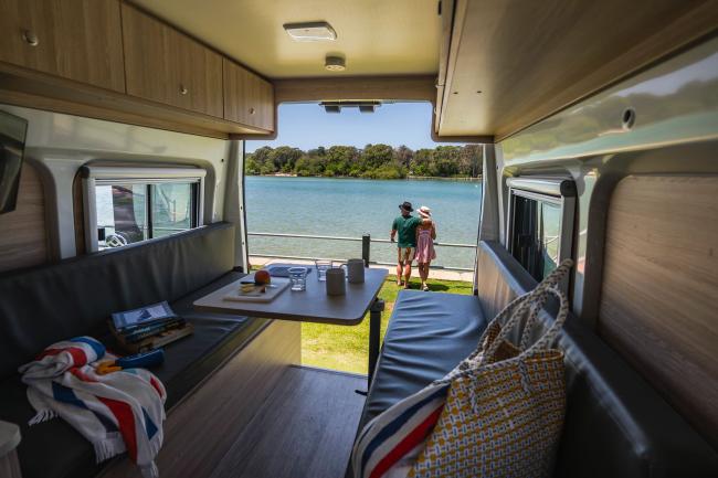 Interior of a campervan looking out the back doors to a couple standing by a river with trees on the far bank.