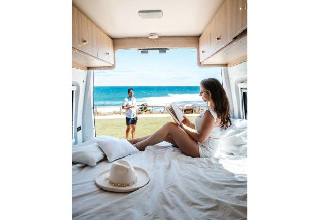 Woman reads on a campervan bed, looking out at a man standing on a grassy lawn overlooking the ocean.