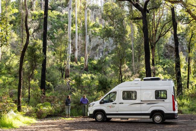 Two people stand near a white Mighty campervan parked in a forest with tall gum trees and rock cliffs.