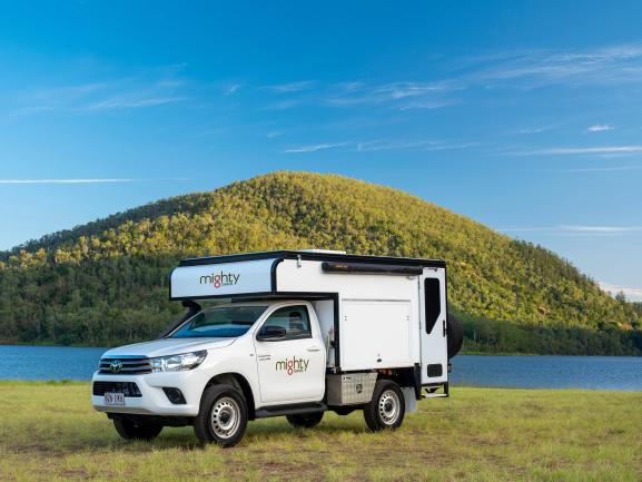 White Mighty 4WD campervan parked on a grassy field beside a lake with a forested hill in the background.