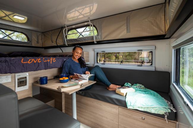 Woman relaxes on a bench seat inside a campervan, reading a book at a table with a bed in the background.