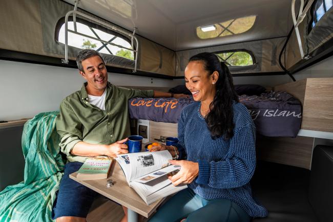 A smiling man and woman sit at a table inside a campervan, reading a book and holding a mug.