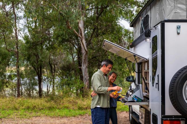 A couple prepares food at a campervan's slide-out external kitchen in a grassy bushland setting.
